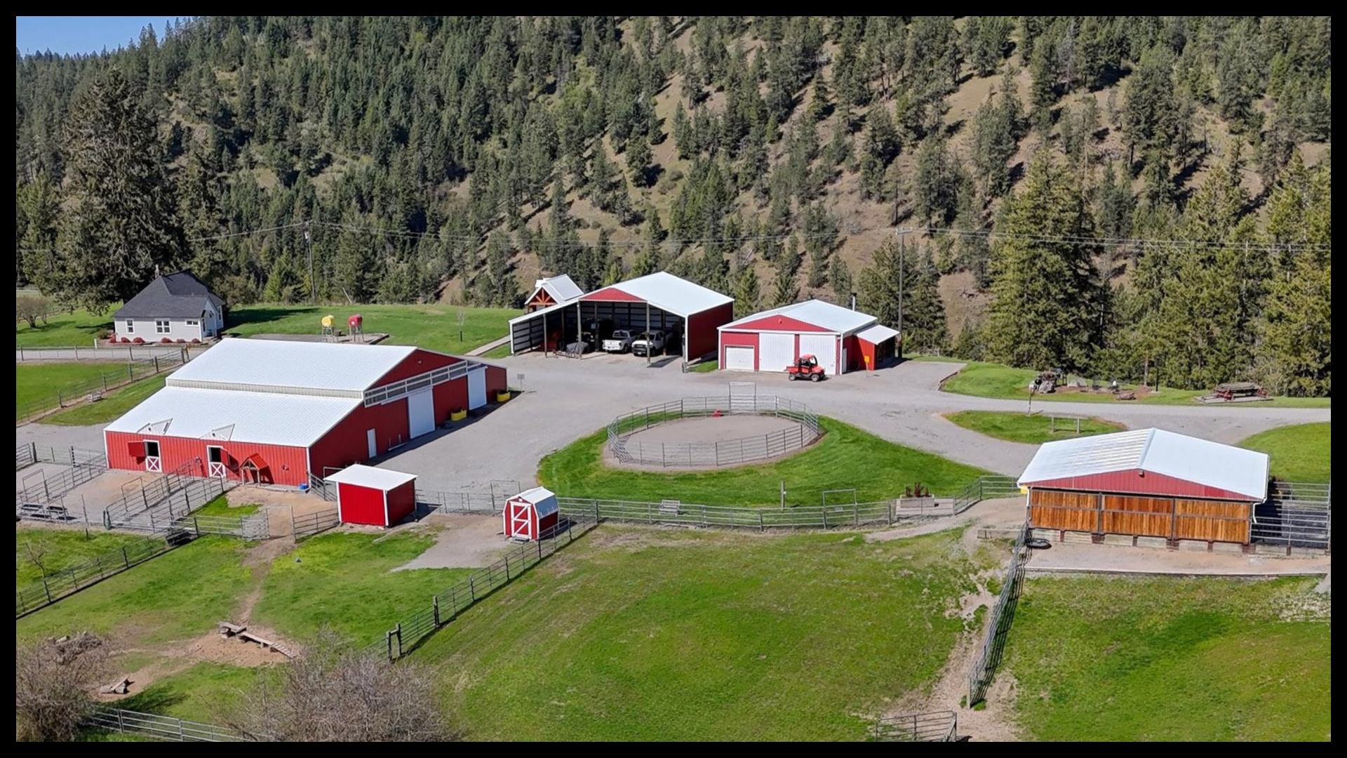 Aerial view of a multi-use ranch property near Coeur d’Alene, Idaho, featuring multiple red barns, a round horse training pen, fenced pastures, and surrounding forested hills.