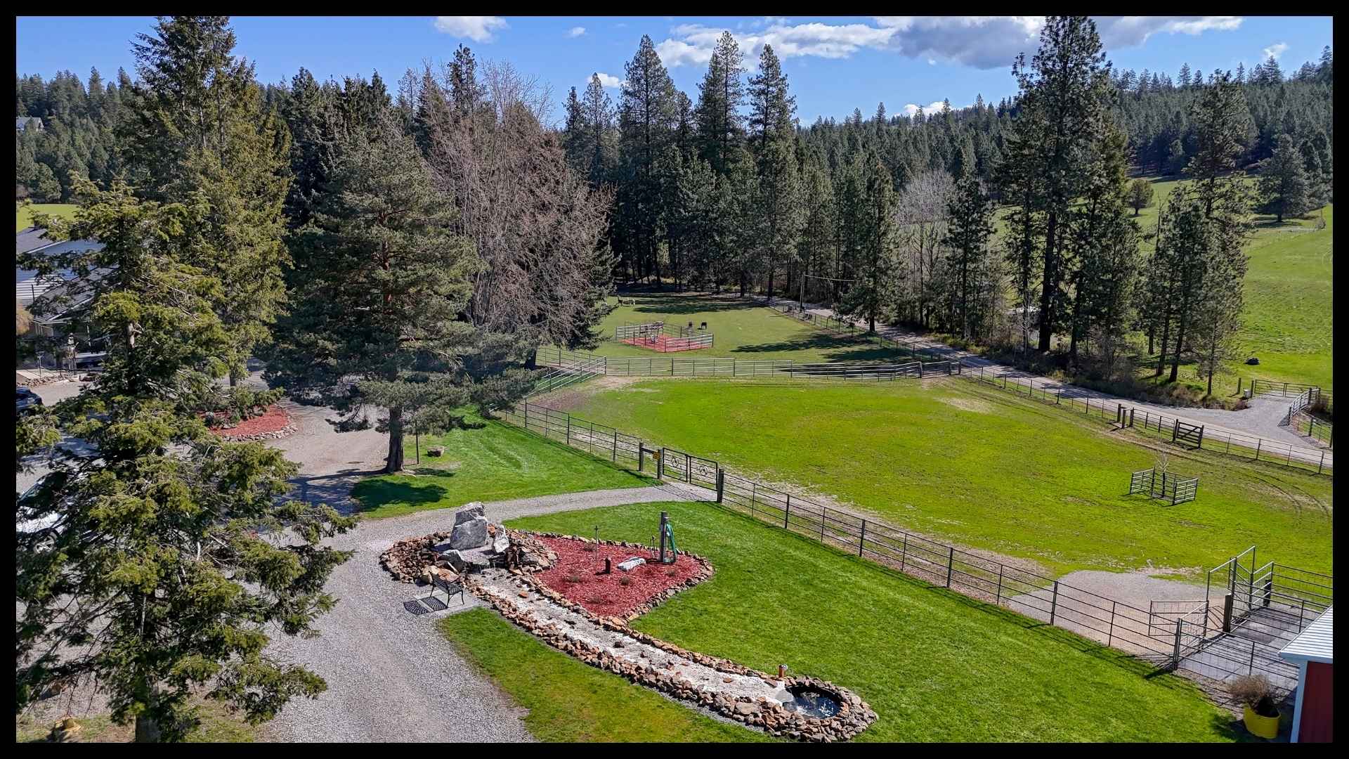 aerial view of a private ranch driveway leading past green pastures, fenced areas, and tall pines with a landscaped rock feature in the foreground