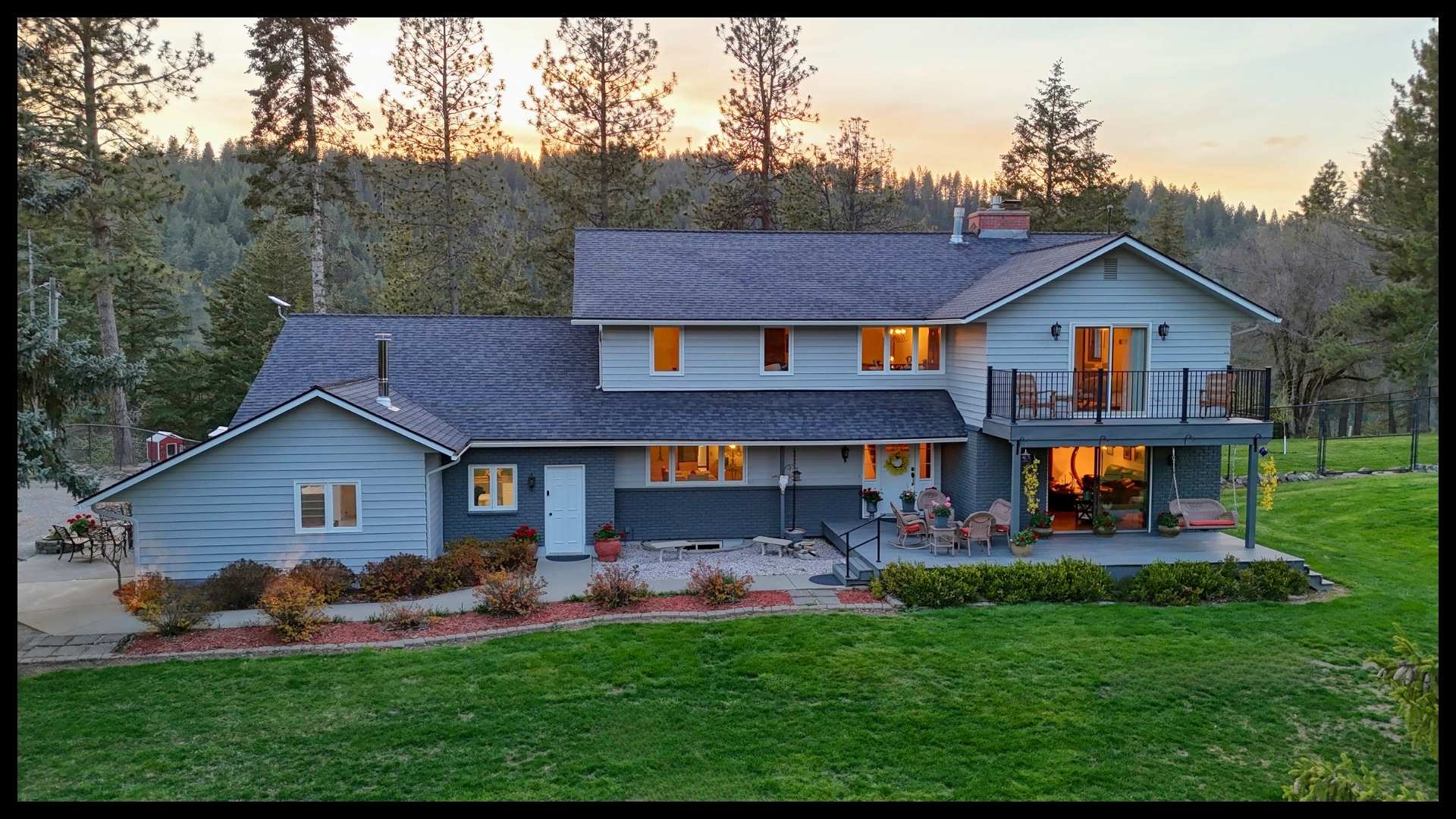 front view of the two-story main home at 2885 S Folsom Ridge Rd, featuring a large balcony, outdoor seating areas, manicured landscaping, and surrounding evergreens at sunset